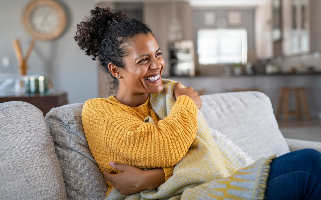 A black woman in a yellow sweater sitting at home on the couch recovering from breast reconstruction surgery
