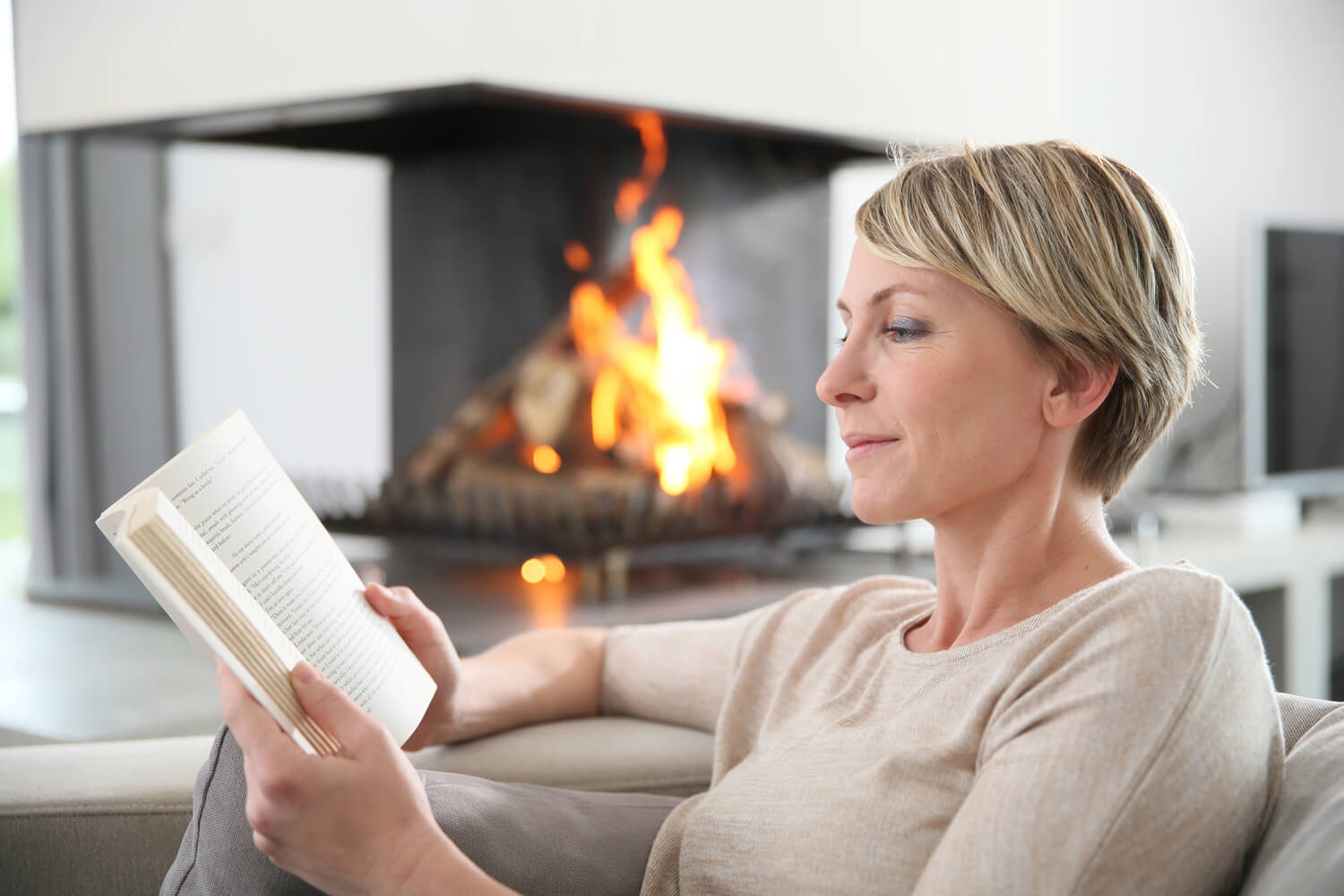 A middle aged blond woman with short hair reading a book next to a fireplace, with no visible signs of vascular surgery.